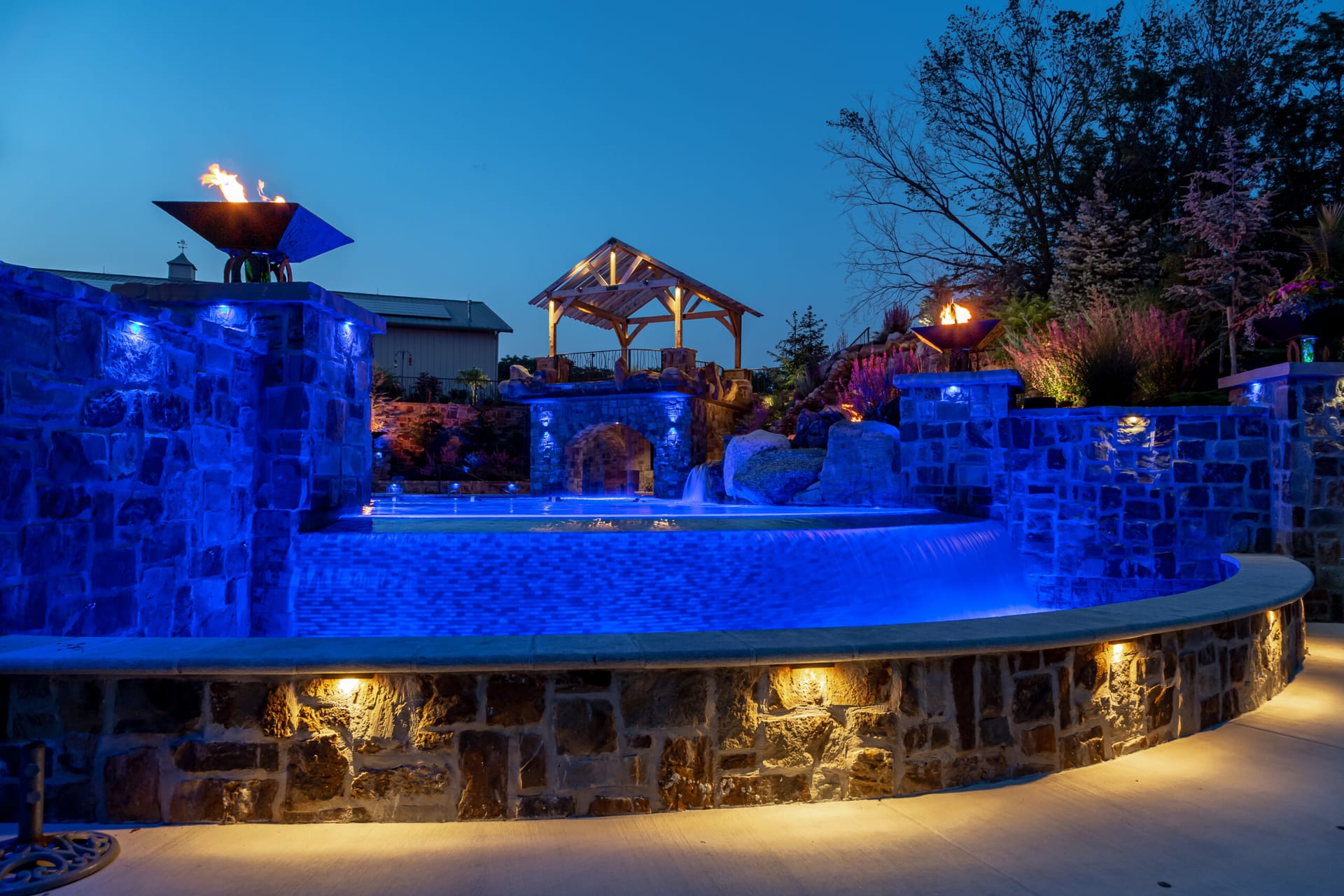 A custom infinity pool lit blue at night with a stone fire feature, built by By The Blade